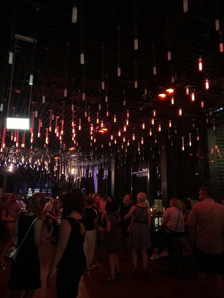 Stimmungsvolle After Work Party in einer modernen Location mit langen, herabhängenden Leuchtröhren in Rot- und Weißtönen, tanzenden Gästen und Barbereich im Hintergrund.
