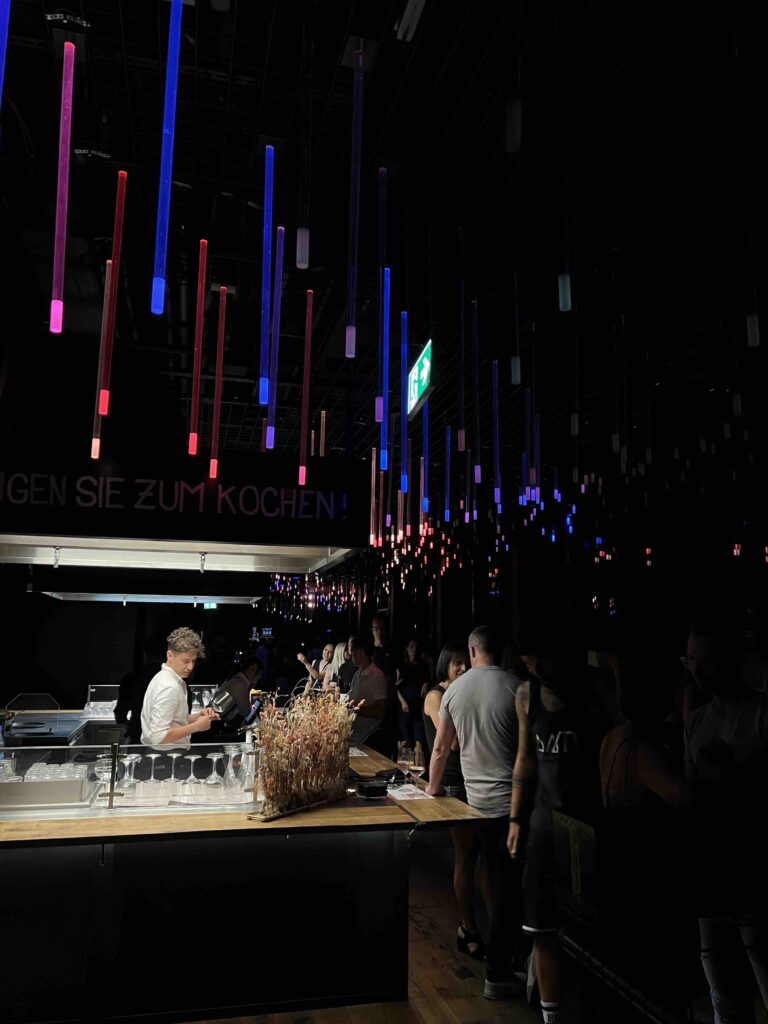 Stylishe After Work Party mit modernen Leuchtröhren in Blau, Rot und Lila, während Gäste an der Bar Getränke bestellen.