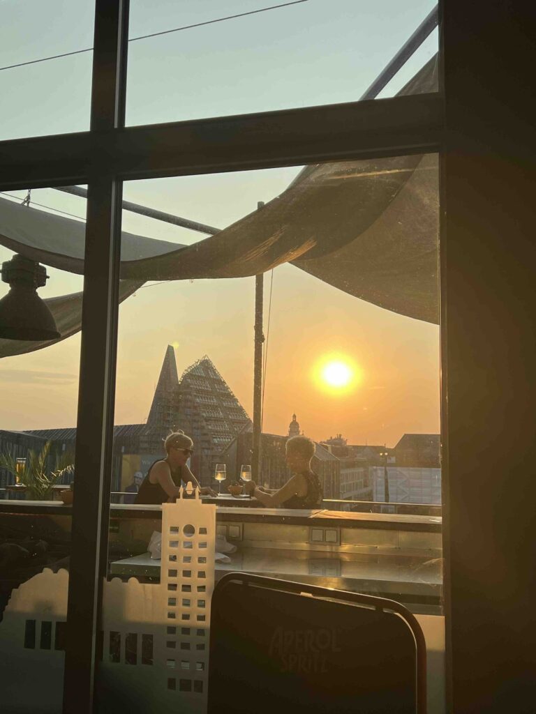 Zwei Frauen sitzen während der After Work Party bei einem Drink auf der Rooftop-Bar und genießen den Sonnenuntergang mit Blick auf den Augustsuplatz von Leipzig.