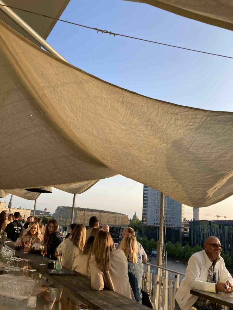 Gesellige After Work Party auf der Rooftop-Bar bei Sonnenuntergang, mit Blick auf Leipzigs Skyline und entspannten Gästen bei Drinks.“