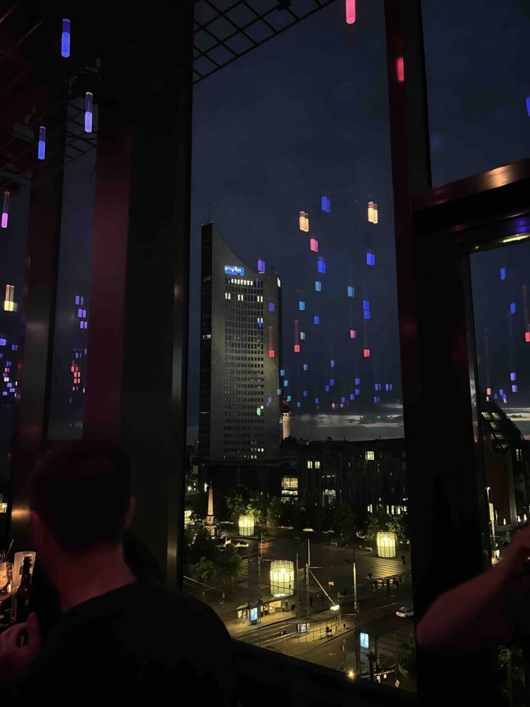 Nachtaufnahme des Uniriesen-Gebäudes in Leipzig, gesehen aus der Rooftop-Bar des FELIX am Augustusplatz – Gäste genießen die After Work Party mit leuchtenden Lichteffekten, urbanem Flair und entspanntem Networking.